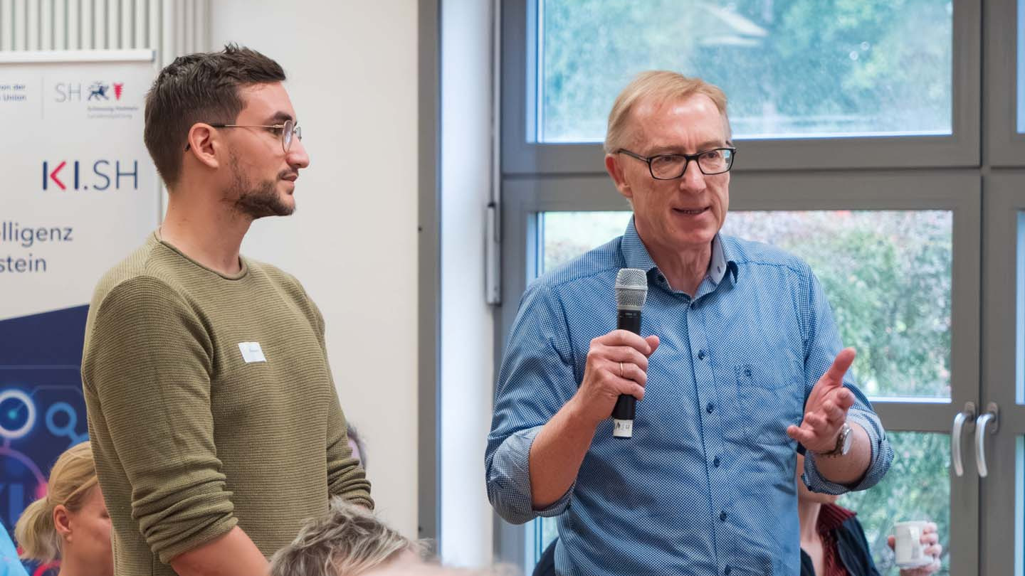 Ein Mann spricht in ein Mikrofon und steht neben einem weiteren Mann